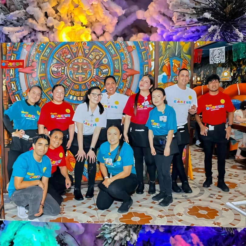 Eleven Fiesta Cancún staff members pose in front of a colorful Aztec calendar mural.