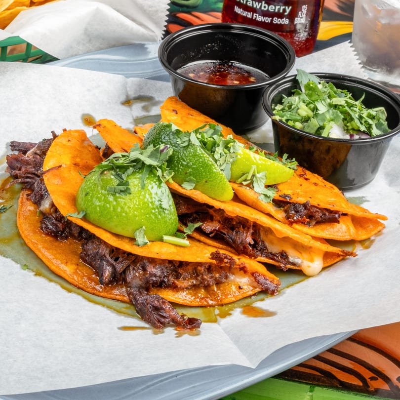 Four birria tacos with limes and cilantro, with dipping sauce and a side of cilantro and onions.