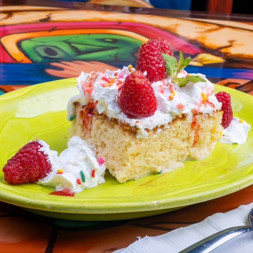 A slice of Tres Leches cake with whipped cream, raspberries, and mint on a lime green plate.