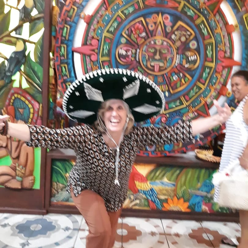 A woman in a sombrero smiles, arms outstretched, in front of colorful Mexican decor.