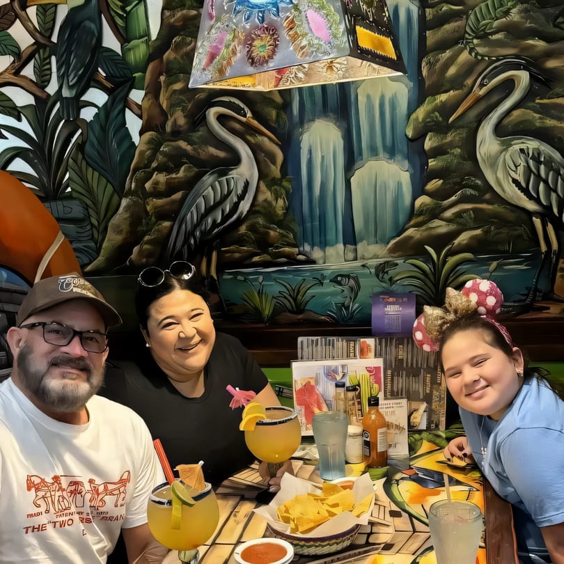 A family of three smiles at their table with drinks and chips at Fiesta Cancún Mexican Restaurant.