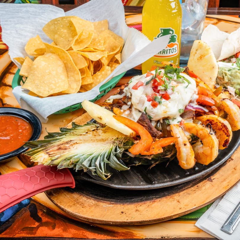 A pineapple filled with shrimp, steak, vegetables, and cheese, served with chips, salsa, and a drink.