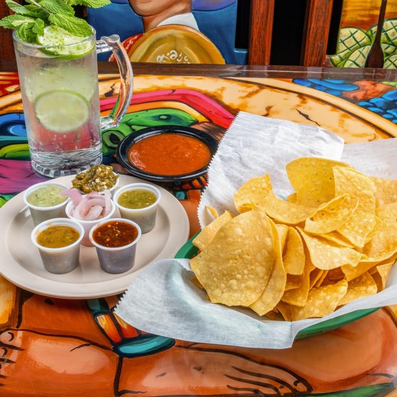 A glass of lime and mint water, a bowl of salsa, and a basket of tortilla chips with various dips.